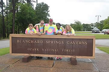 Picture of students and staff in yellow blue pink swirled tye dyed shirts stand behind Blanchard Springs Caverns Sign