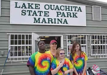 Students and staff in rainbow tye dyed shirts stand in front of Lake Ouachita State Park Marina Sign