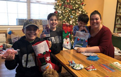 Three young boys and an adult woman gather around a table covered in holiday goodies and smile at the camera. In the background is a beautifully decorated Christmas tree.